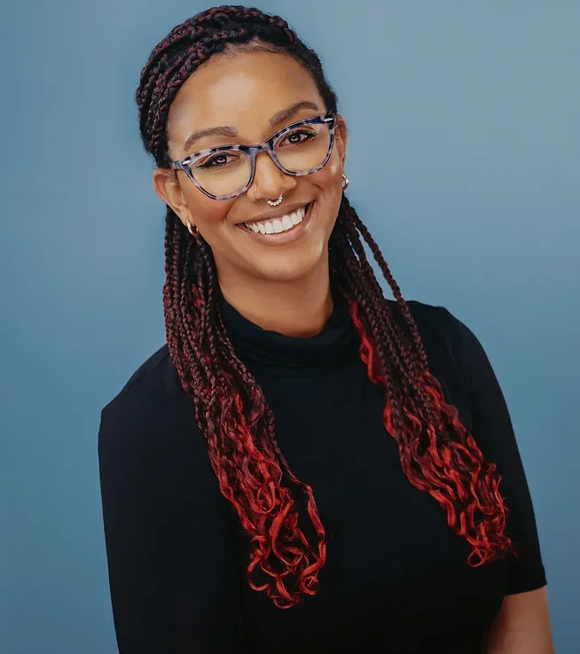 Portrait of Jasmine Patterson smiling, wearing glasses and a black top, with long red-tipped braids, against a blue backdrop